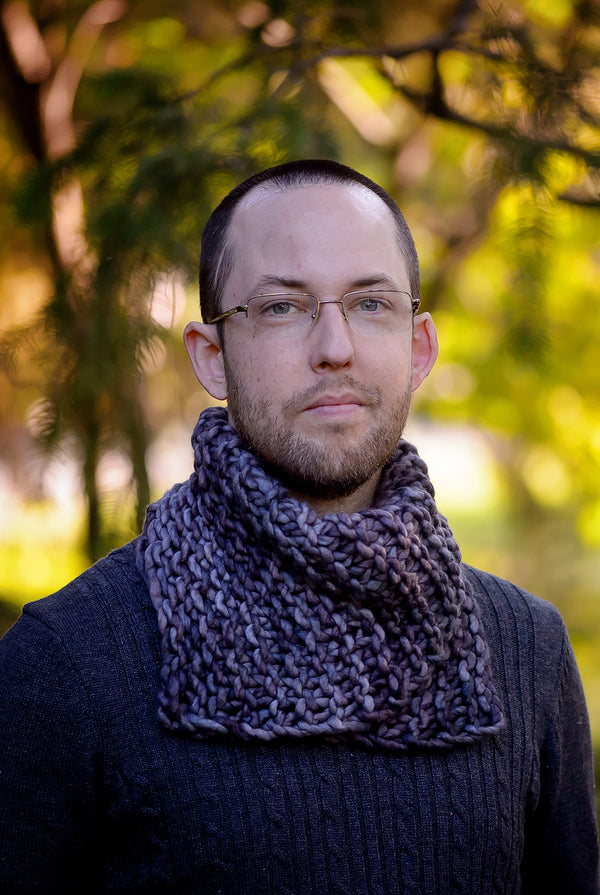 Our Asymmetrical Cowl in the colour Plomo shown on a young man in a forest.