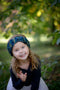 A little girl reaches towards the camera while playing in the forest while wearing a Little Twist Headband. Shown in Verde Azul.
