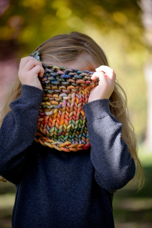 A small child pulling a cowl over her face in the woods. Shown in Arco Iris
