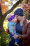A mother holds her child while wearing our Snowball Fight Hat in the woods. Shown in Plomo.