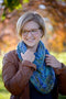 The Triangulate Cowl in colour Indicita, is worn by a woman in a brown leather jacket. Autumn coloured trees in the background.